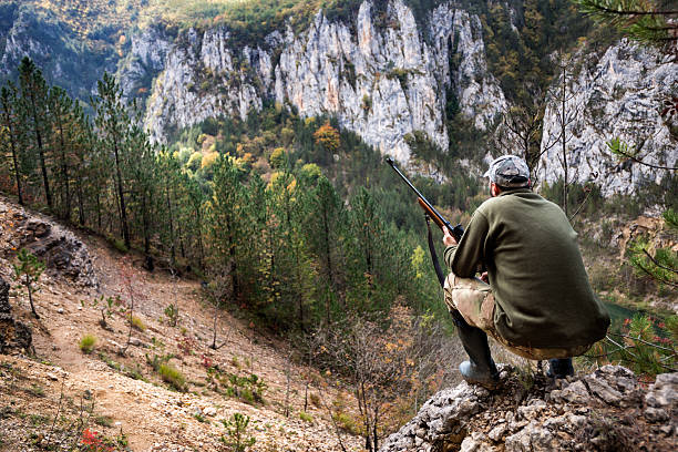 Portrait of hunter with rifle crouching on top of the hill and stalking his prey.